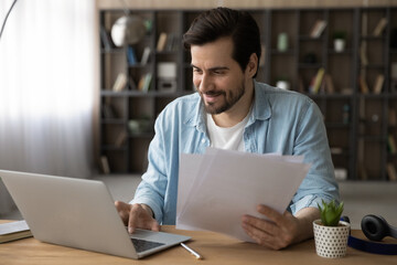 Happy handsome young businessman manager holding paper documents in hands, analyzing sales data or research report statistics using computer software application, working distantly at home office.