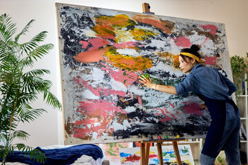Inspired female artist working on a large modern abstract oil painting, applying paint on canvas with fingers, standing in home studio workshop