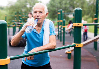 Obraz premium old peppy pensioner man resting and drinking water on a sports equipped playground