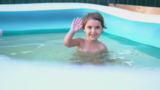 Happy little girl having fun inside a swimming pool outdoors in summer, waving hand and she is pleased.