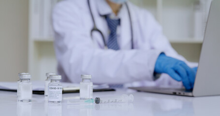 Covid-19 vaccine on the desk with hands of doctor typing on notebook computer for register the...