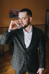 Stylish, handsome, smiling male businessman in a jacket drinks whiskey from a glass. Preparing for the meeting.