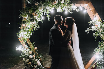 Stylish groom in a vest and a beautiful bride in a white long dress hug near a wooden arch,...