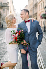 Close up portrait of loving couple, 50-aged handsome man in suit and bow tie, standing with his pretty woman in white dress with flower bouquet, on the background of old city