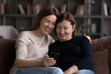 Happy mature 60s mother and grownup daughter using mobile phone together, making video call, watching pictures, browsing internet, social media, having fun, hugging, enjoying meeting and leisure time