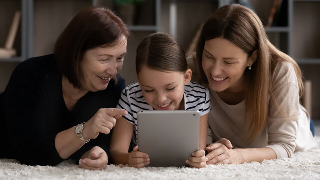 Happy three female generations of family, girl, mom, grandma relaxing at home together, lying on heating comfortable floor, using tablet, watching online video on device, making call
