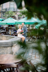 Romantic beautiful middle-aged couple standing in a city square alongside the fountain. View through the green leaves