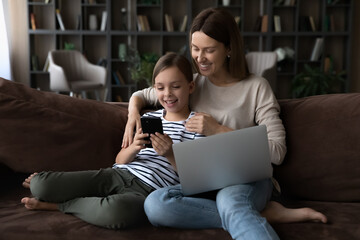 Happy gen Z girl and mom relaxing on couch with gadgets. Mother and little daughter kid using parental control app on smartphone, playing online game, watching video on laptop, browsing internet