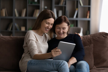 Happy grownup daughter showing content on tablet to mature mother. Two family generations women with digital computer gadget resting on couch together, watching videos, making video call