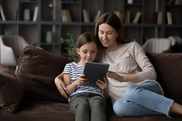 Happy relaxed mom and gen Z daughter kid with gadget enjoying leisure at home together, resting on sofa, using app or service on tablet, watching movie, video, reading book, browsing internet