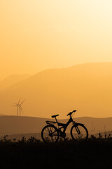 Fototapeta premium Mountains bike silhouette and setting sun, orang sunset, wind turbines and mountains on background.