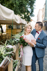 Happy mature couple in fashionable clothes, posing near flower pots decorations at outdoors city cafe on summer day in old ancient European city