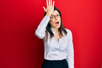 Young hispanic woman wearing business shirt and glasses surprised with hand on head for mistake,...