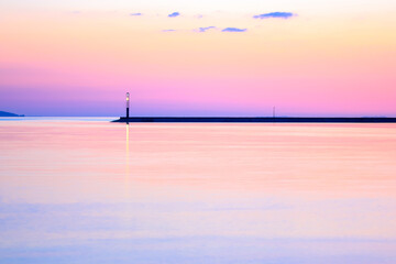 A concrete breakwater on a horizon. A beautiful seascape. A purple sky and reflection in the water water without waves at sunset. Anti-wave protection. Lighthouse glowing in a distance. Twilight hour.