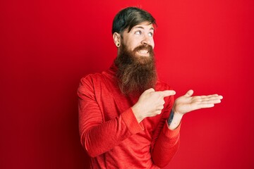 Redhead man with long beard pointing open palm with finger smiling looking to the side and staring away thinking.