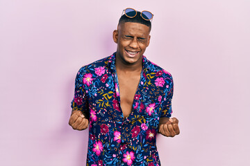 Fototapeta premium Young black man wearing hawaiian shirt and sunglasses very happy and excited doing winner gesture with arms raised, smiling and screaming for success. celebration concept.
