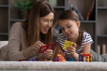 Obraz na płótnie Canvas Cute girl and happy mom engaged in learning game together, playing construction toys, building blocks, connecting pieces, plastic details, relaxing on clean carpet on heating floor. Family playtime