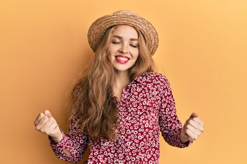 Beautiful blonde caucasian woman wearing summer hat very happy and excited doing winner gesture with arms raised, smiling and screaming for success. celebration concept.