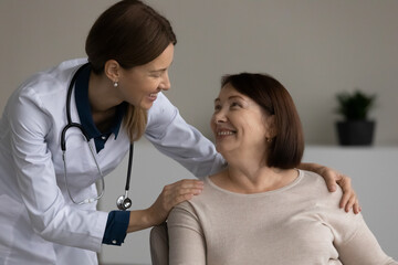 Happy positive medical professional, doctor, nurse giving support and help to senior female patient at appointment, touching shoulders. Old woman visiting, consulting young physician in office