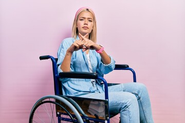 Beautiful blonde woman sitting on wheelchair rejection expression crossing fingers doing negative...