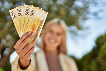 Young blonde girl smiling happy holding norway krone banknotes at the city.