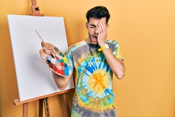 Young hispanic man standing drawing with palette by painter easel stand yawning tired covering half...