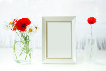 mock up white photo frame on the table by the window and two vases with daisies and poppies..