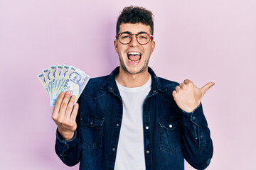 Young hispanic man holding 100 romanian leu banknotes pointing thumb up to the side smiling happy with open mouth