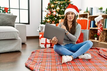 Young latin woman using laptop sitting by christmas tree pointing with hand finger to the side showing advertisement, serious and calm face