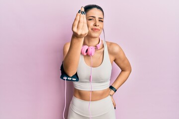 Young hispanic woman wearing gym clothes and using headphones doing italian gesture with hand and fingers confident expression