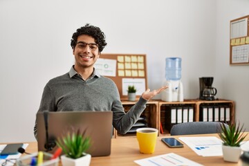 Fototapeta premium Young hispanic man wearing business style sitting on desk at office smiling cheerful presenting and pointing with palm of hand looking at the camera.
