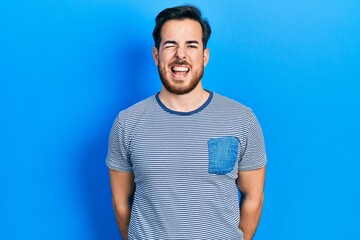 Handsome caucasian man with beard wearing casual striped t shirt winking looking at the camera with sexy expression, cheerful and happy face.