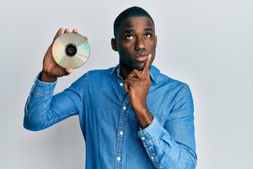 Young african american man holding compact disc serious face thinking about question with hand on...