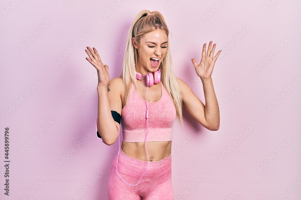 Poster young beautiful caucasian woman wearing gym clothes and using headphones celebrating mad and crazy f