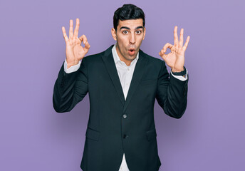 Handsome hispanic man wearing business clothes looking surprised and shocked doing ok approval symbol with fingers. crazy expression