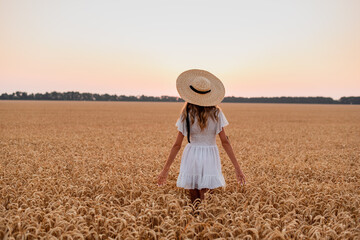 the girl meets the dawn on a wheat field. She touches the spikelets with her hands and walks along the field. On her head is a straw hat with ribbons