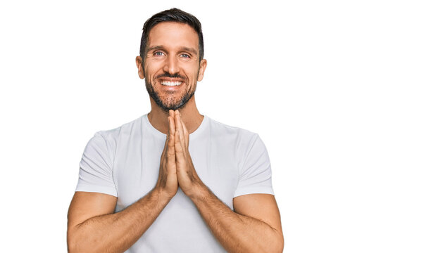 Handsome man with beard wearing casual white t shirt praying with hands together asking for forgiveness smiling confident.