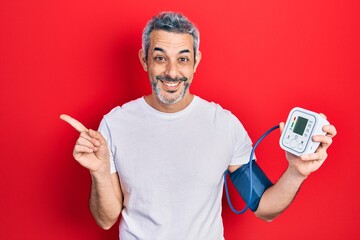 Handsome middle age man with grey hair using blood pressure monitor smiling happy pointing with...