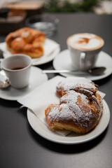Traditional Italian breakfast. Fresh croissants with cream and sugar powder, espresso coffee and cappuccino on a black wooden table in Milan, Lombardy, Italy. European food and pastry.