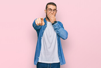 Young caucasian man wearing casual clothes laughing at you, pointing finger to the camera with hand over mouth, shame expression