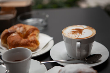 Traditional Italian breakfast. Fresh croissants with cream and sugar powder, espresso coffee and cappuccino on a black wooden table in Milan, Lombardy, Italy. European food and pastry.