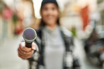 Young hispanic woman smiling happy using microphone at the city. - obrazy, fototapety, plakaty