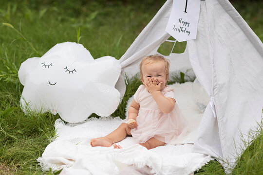 Portrait of cute caucasian baby infant with blue eyes and blonde hair. Beautiful photo session set up for baby girl celebrating one year old birthday, outdoor at summer with white wigwam, blanket