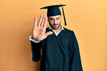 Young hispanic man wearing graduation cap and ceremony robe doing stop sing with palm of the hand....