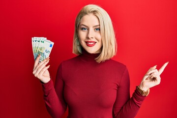 Young blonde woman holding bunch of 5 euro banknotes smiling happy pointing with hand and finger to the side