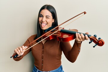 Young brunette woman playing violin sticking tongue out happy with funny expression.