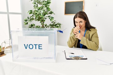 Young brunette woman sitting at election table with voting ballot smelling something stinky and...
