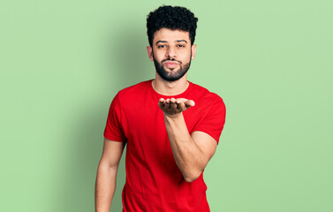 Young arab man with beard wearing casual red t shirt looking at the camera blowing a kiss with hand on air being lovely and sexy. love expression.