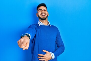 Young hispanic man with beard wearing casual blue sweater laughing at you, pointing finger to the camera with hand over body, shame expression