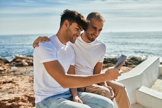 Young gay couple smiling happy using smartphone at the beach.